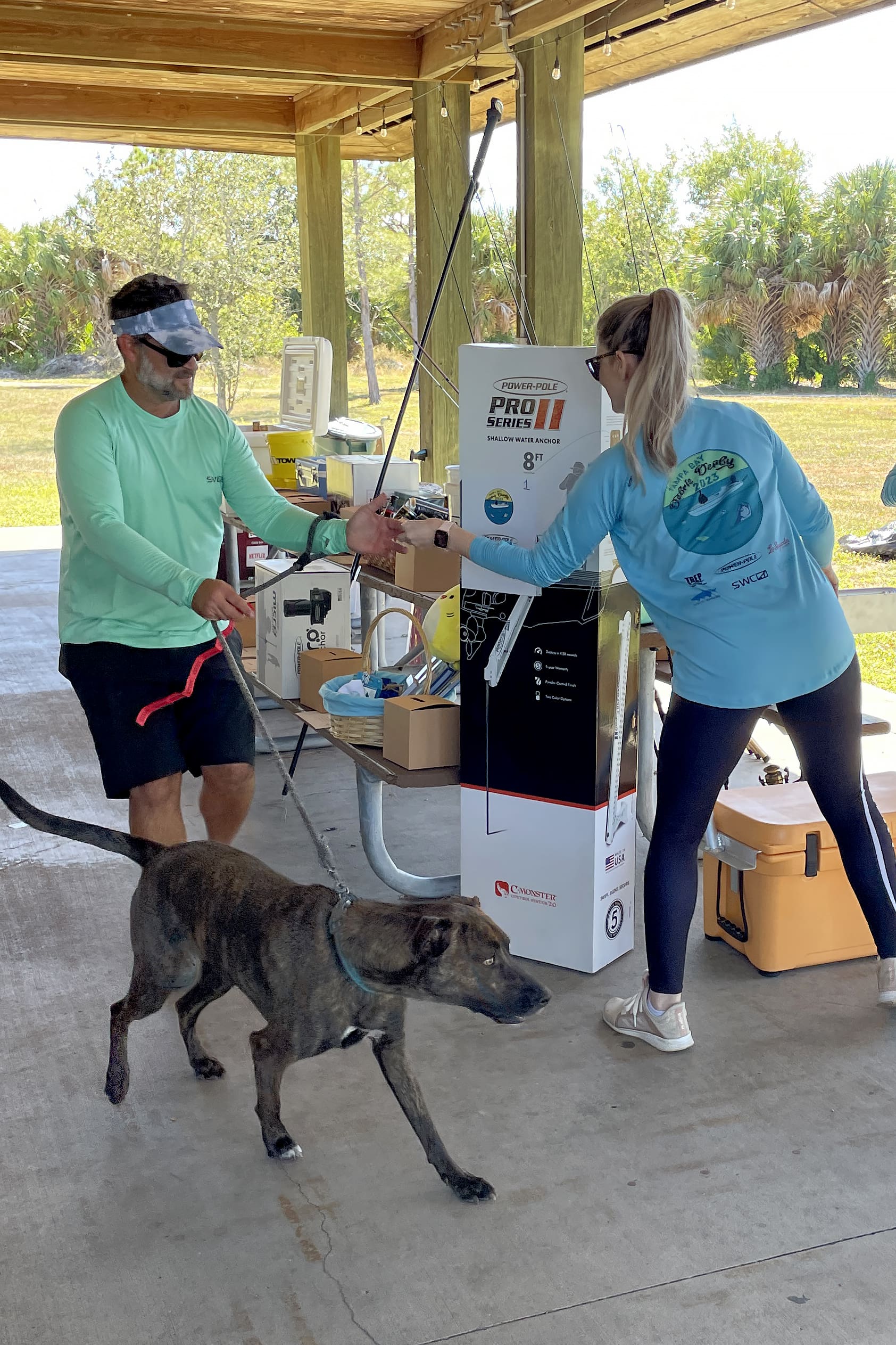 A woman hands a fishing pole to a man holding a dog on a leash.