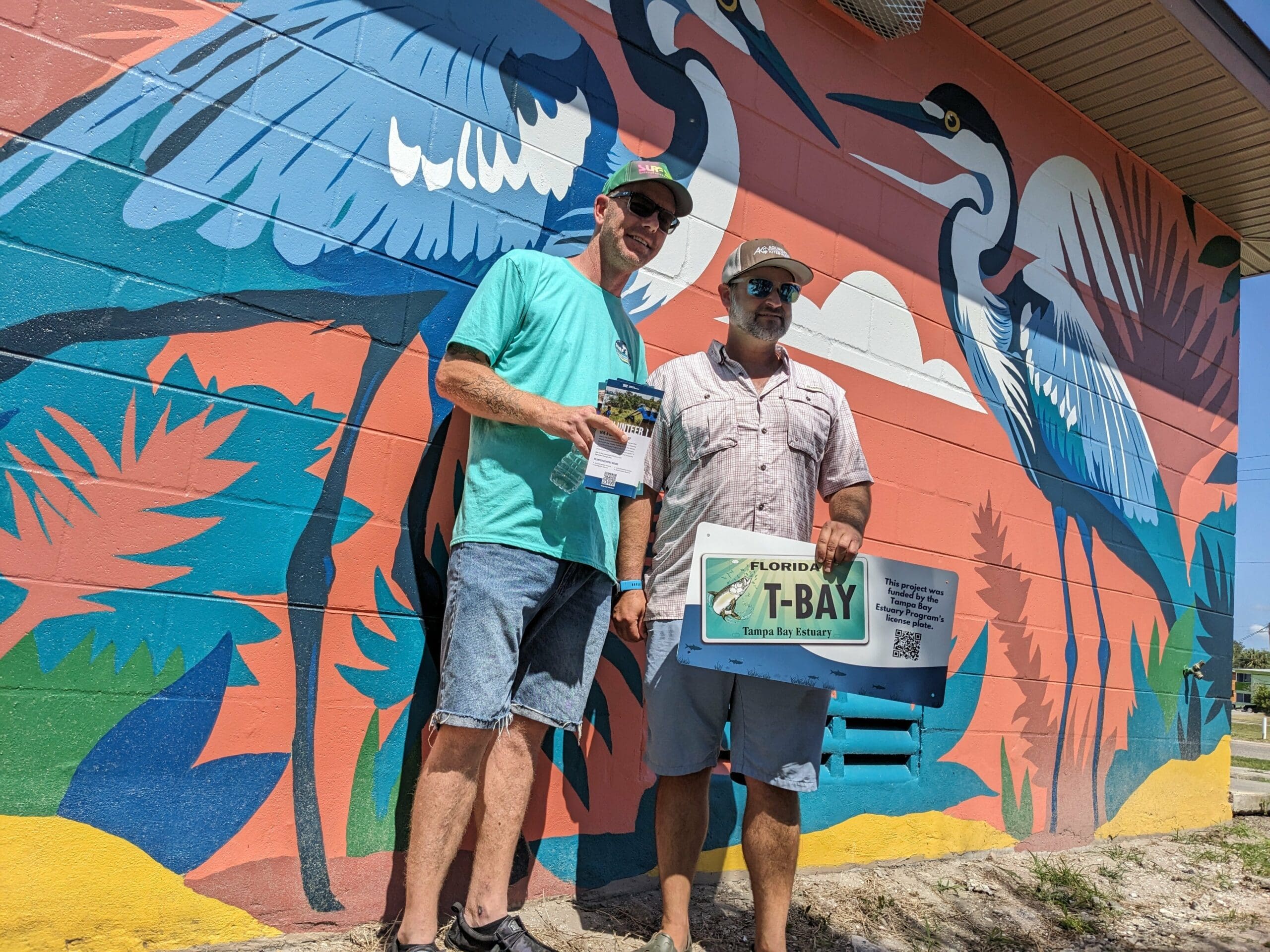 two mean stand in the sunshine in front of a new mural showing great blue herons while holding a bay mini-grant sign